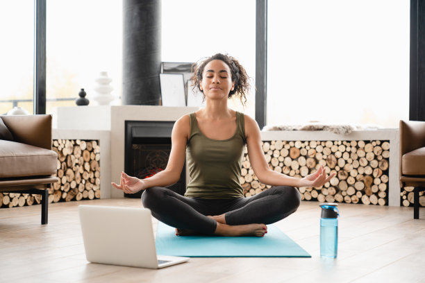 年轻的非洲瑜伽女运动员穿着运动服，在健身垫上冥想莲花位，在家里的笔记本电脑上观看在线培训教程，感觉很放松图片下载