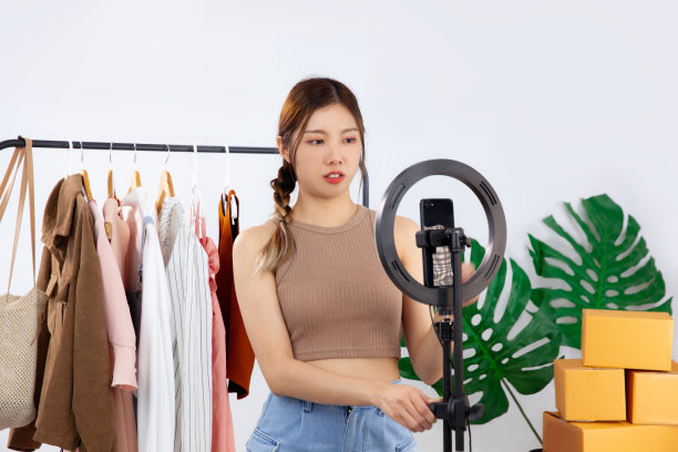 年轻的亚洲妇女在棕色背心设置照明和智能手机直播蒸汽卖她的衣服在家里。图片下载