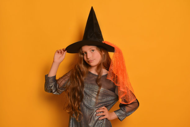 可爱的小女孩在万圣节女巫服装拿着黑色和橙色的帽子，手放在腰上，看着黄色背景的相机。图片下载