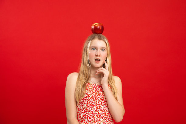 睁开眼睛的少女站在苹果躺在头上，空的红色背景，自由复制空间。聪明的孩子穿着印花连衣裙，带着惊讶的情绪。思想，研究，震撼，科学符号图片下载