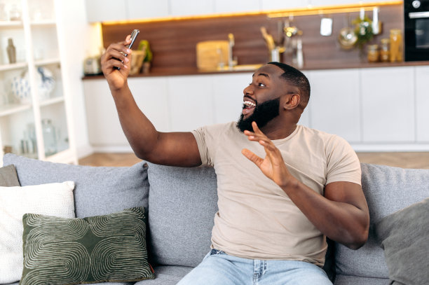 快乐的非裔美国年轻时尚男子坐在客厅的沙发上，使用他的智能手机，通话视频电话。积极的黑皮肤视频博主，用自拍手机，录视频图片下载