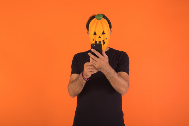 戴着南瓜面具庆祝万圣节的人，看着手机，橙色背景。庆祝、万灵节和万圣节概念图片下载