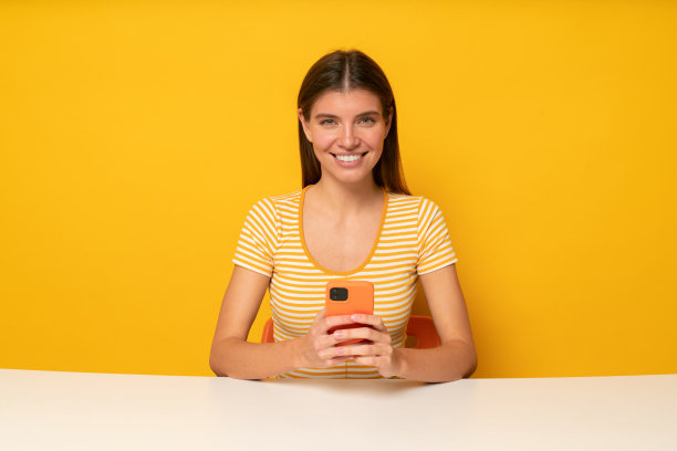 女老师坐在桌子前，手里拿着手机，看着黄色背景的相机图片下载