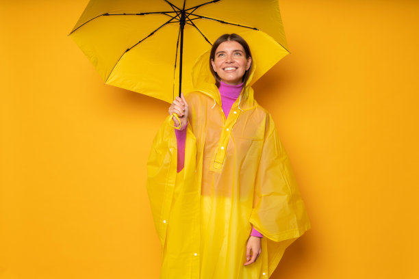 身穿黄色防水雨衣的女子站在伞下，在工作室背景上仰望天空图片下载