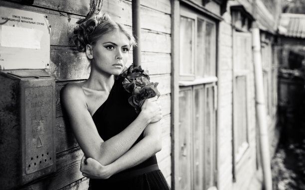 美丽的年轻女子穿着黑色的连衣裙，戴着红色的花朵，正坐在破旧的木头建筑的危险上图片下载