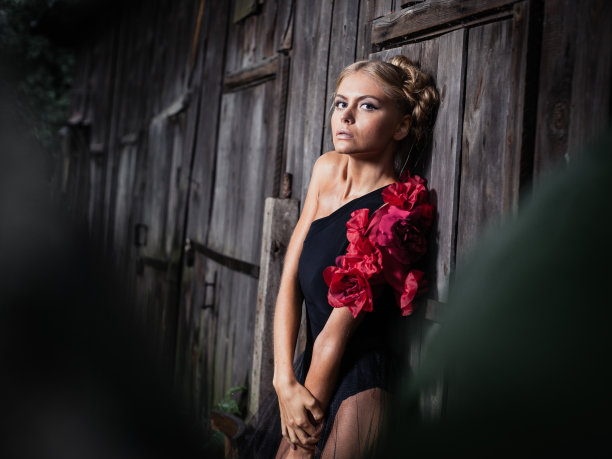 美丽的年轻女子穿着黑色连衣裙，戴着红花，站在破旧的木墙旁图片下载
