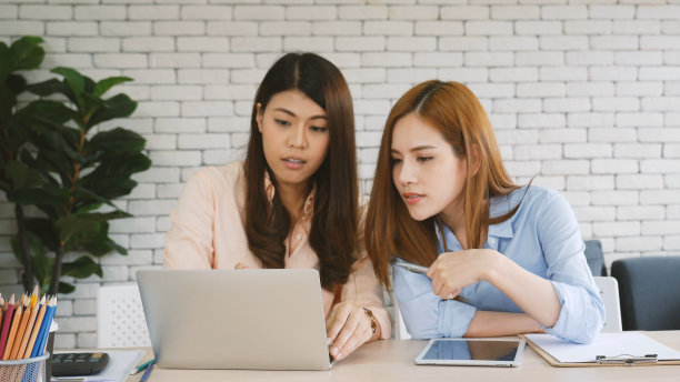 两个女商人在公司办公桌旁用笔记本电脑开会。两个年轻的女性自由职业阅读金融图表策划分析市场数据。亚洲人团队工作办公室公司。图片下载