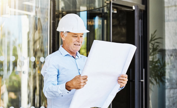 戴着安全帽的中年建筑老板、土木工程师或建筑师站在建筑物的玻璃入口外检查平面图图片下载
