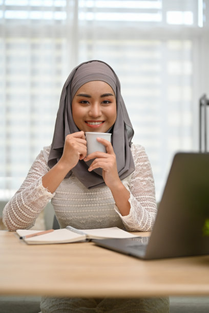 美丽的亚洲穆斯林妇女戴着头巾，一边喝咖啡一边在办公桌前工作。图片下载