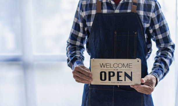 一个穿着围裙的店主举着一个开着的牌子。女服务员站在咖啡馆的入口处准备食物。图片下载