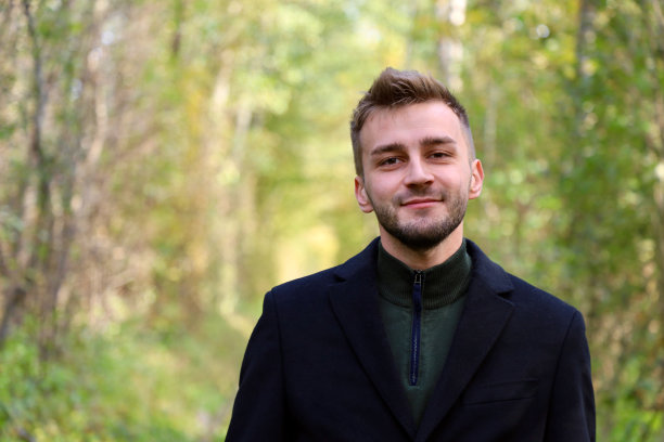 森林里穿黑大衣的帅哥。可爱的家伙留着胡茬，留着模特发型，穿着外套站在森林里摆姿势。图片下载