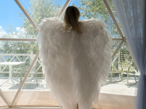 一名身穿白色运动紧身衣、长着天使翅膀的金发女子站在夏季招待所的全景窗口前图片下载