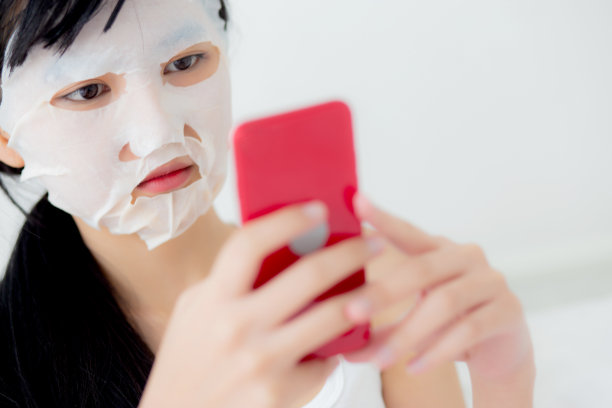 美丽的年轻亚洲女性与床单面膜坐在床上看智能手机在卧室，美丽的女孩敷面膜的皱纹在家里，皮肤护理，健康和健康。图片下载