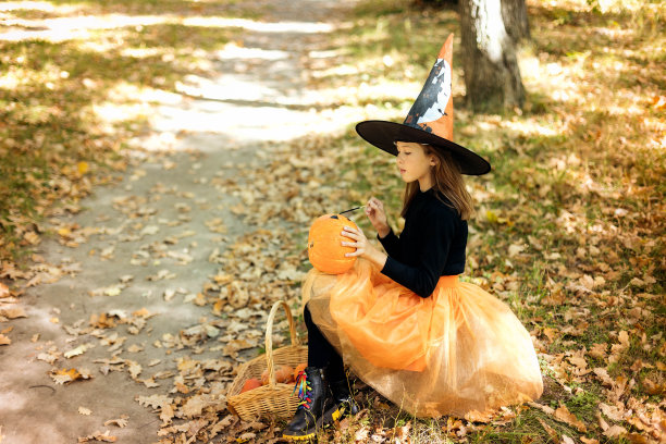 一个迷人的女孩穿着女巫服装坐在树林里，用魔杖在南瓜上画画，万圣节图片下载