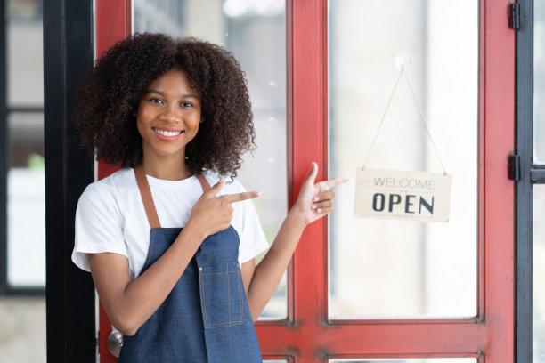 一个微笑着的年轻非裔美国咖啡师的肖像，斜倚在一家时髦的咖啡馆的门上，双臂交叉图片下载