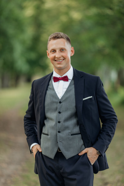 画像年轻微笑男子新郎短发，穿着蓝色西装，灰色背心，红色领结，站在公园的夏天。图片下载