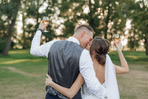 年轻男子新郎亲吻拥抱年轻美丽的女子新娘，举起手拿酒杯。婚礼,庆典。图片下载