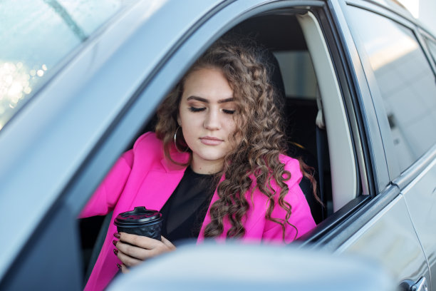 年轻女子开车。画像中的卷发女人在车里望着窗外，喝着热咖啡图片下载