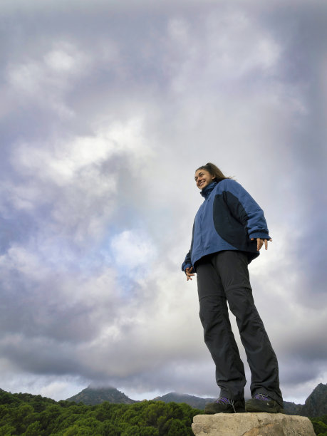 在多云的天空下，站在岩石上的女孩看着地平线微笑。图片下载