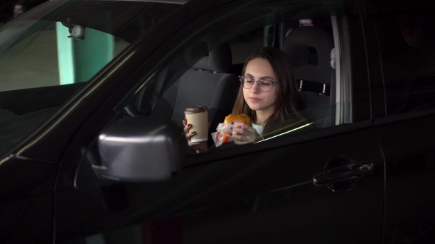 女商人在车里吃午饭，吃得很快。一个戴眼镜、穿西装的体面女人吃着芝士汉堡，喝着咖啡。图片下载