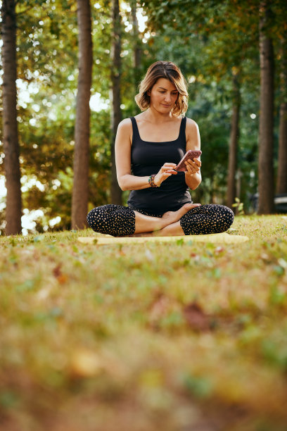 一名瑜伽女子在森林里摆着莲花式坐着，用着智能手机。图片下载