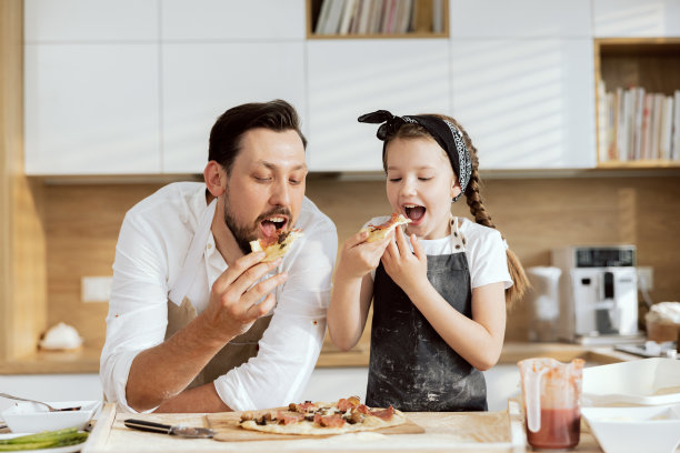 快乐的父亲和快乐的孩子穿着围裙，在现代厨房品尝美味可口的自制披萨。高兴的父母看着微笑的女儿。家人花时间在一起玩图片下载