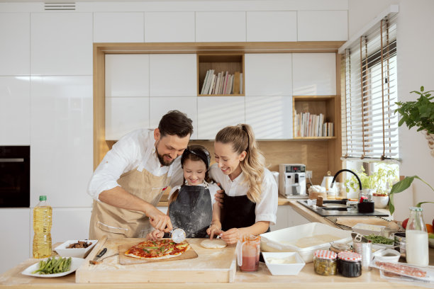 高兴的孩子，孩子，女儿，切着自制披萨，穿着围裙。幸福的父母和家人在有大窗户的现代厨房共度时光图片下载