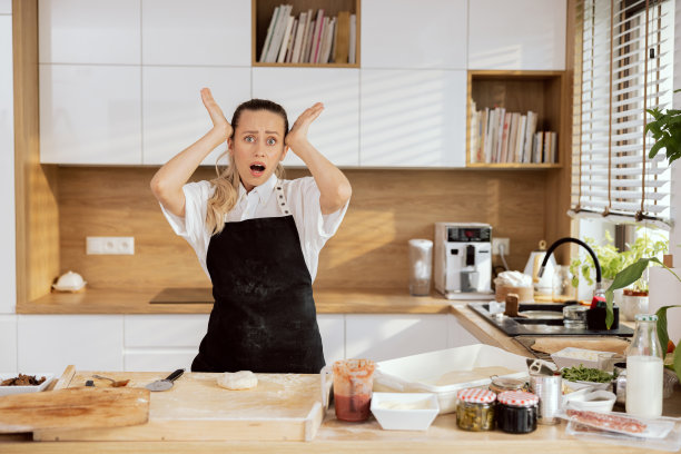 困惑震惊的年轻女子女厨师穿着围裙，手在头附近，看着相机，嘴巴。桌上的披萨配料番茄酱橄榄芦笋黄油面粉鸡蛋烘焙图片下载