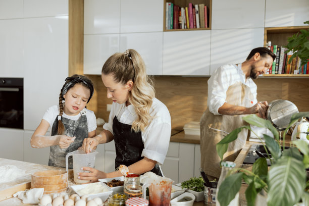 前景中可爱的母女用勺子在量杯里倒面粉。在前景年轻的父亲穿着围裙在水槽里洗碗。幸福的家庭在现代厨房有乐趣图片下载