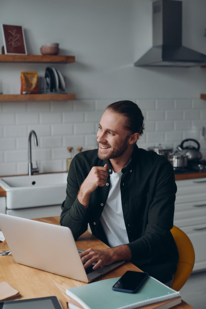 英俊的年轻人使用笔记本电脑，微笑着在家里的厨房柜台工作图片下载
