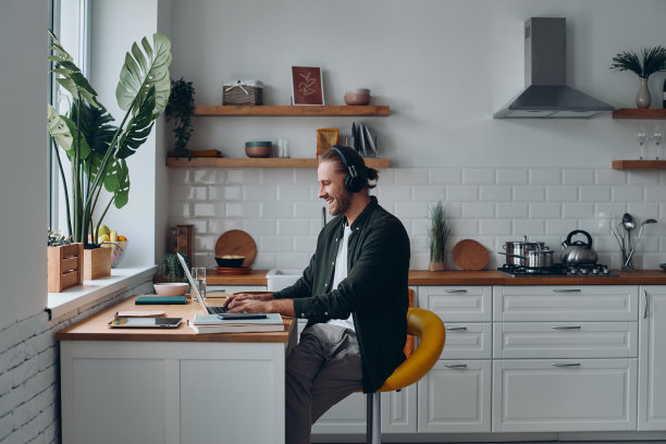 快乐的年轻人戴着耳机坐在家里的厨房柜台上用笔记本电脑工作图片下载