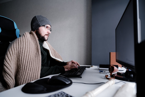 年轻人戴着眼镜，戴着暖和的帽子，裹着毯子，在电脑前工作。房间里很冷。关掉暖气和煤气，降低房间温度的概念。侧视图图片下载