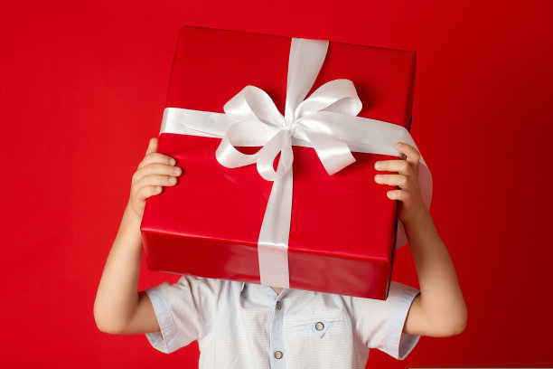 小男孩手里拿着一个大大的红色礼品盒，上面有白色的蝴蝶结，他藏在礼品盒后面图片下载