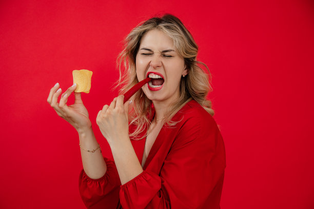 欢快，高兴的金发女人品尝垃圾辣辣椒和美味的咸薯片在红色的背景。热的辣的食物图片下载