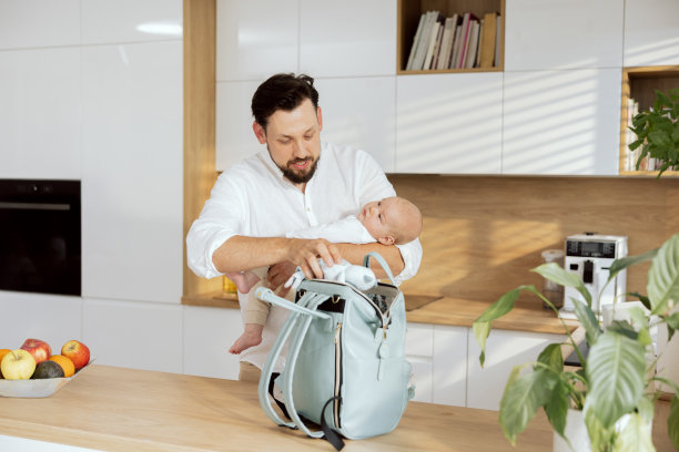 快乐的父亲背着背包，把玩具混合物放进去喂孩子。英俊的父亲双手抱着婴儿站在现代厨房的木桌前，和年轻的母亲一起散步图片下载