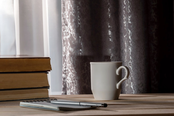 一杯咖啡或茶放在桌子上，笔记本和一堆书靠近窗户有窗帘。副本的空间。图片下载