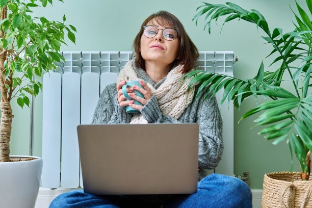 穿着暖和衣服的妇女坐在暖气散热器附近，使用笔记本电脑图片下载