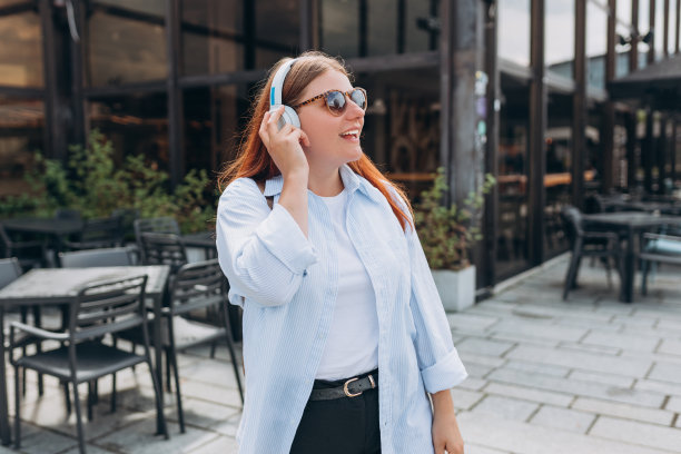 可爱快乐的年轻女子戴着太阳镜，在城市背景下通过无线耳机听音乐。喜欢音乐的人。生活方式的概念。走在城市街道上的女孩图片下载