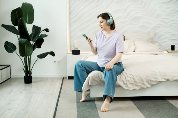 微笑的成年妇女坐在床上在卧室戴着耳机，听音乐图片下载