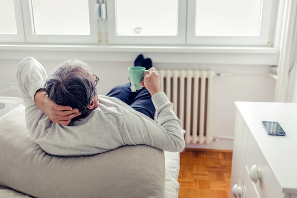 从肩膀上看，一位年长的男子喝着热茶，把脚放在暖气片上取暖，穿着羊毛袜子，坐在窗边，呆在家里的多雨的冬天。图片下载