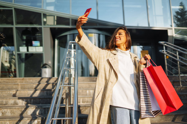 年轻女子站在路边拿着购物袋和使用手机。图片下载