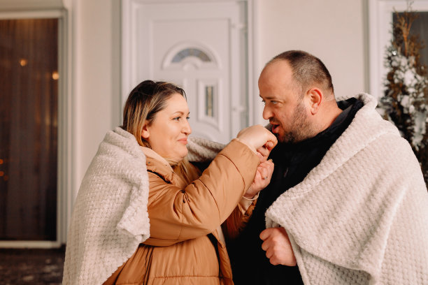 特写照片的女人和男人站在前面的房子在晚上穿着温暖的衣服盖着毯子和男人温暖妻子的手与他的呼吸。图片下载