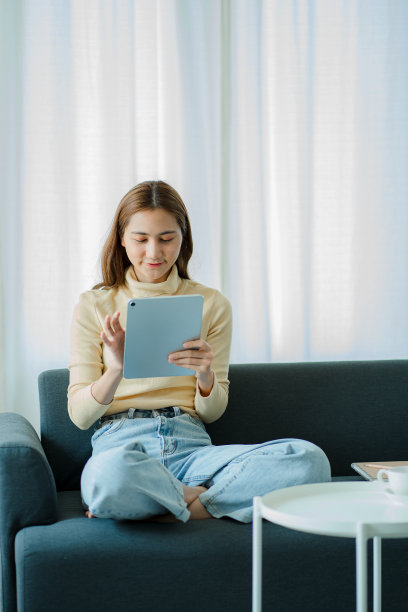 快乐的亚洲妇女拿着电脑平板电脑技术坐在家里的沙发上使用应用程序，网上购物，阅读新闻，上网冲浪。图片下载