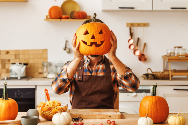 一名穿着围裙的男子在厨房雕刻橙色万圣节大南瓜时，用南瓜遮住了他的头图片下载