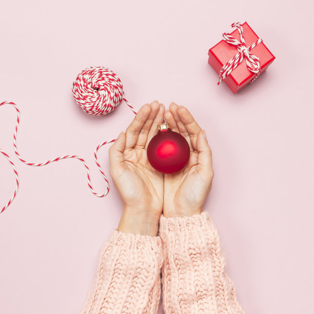 女性手在粉红色的针织毛衣拿着一个红色的圣诞球，礼品盒在粉红色的背景平铺风格俯视图。庆典、节日、圣诞节、新年、装饰物件图片下载