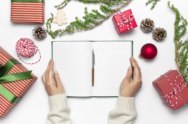穿着针织毛衣的女人们正用钢笔在干净的笔记本上写着新年计划，礼品盒、杉树树枝在白色的桌子上平铺着。圣诞策划概念2019年的节日装饰图片下载
