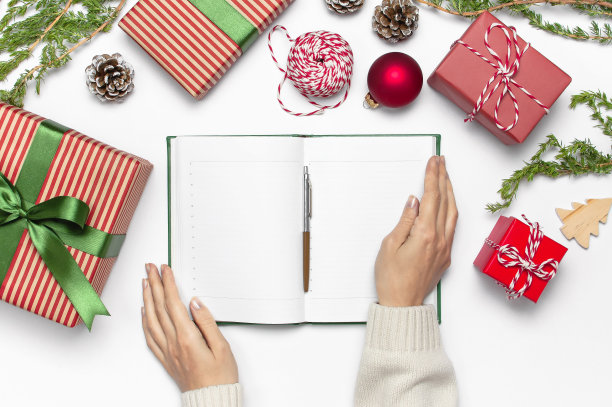 穿着针织毛衣的女人们正用钢笔在干净的笔记本上写着新年计划，礼品盒、杉树树枝在白色的桌子上平铺着。圣诞策划概念2019年的节日装饰图片下载