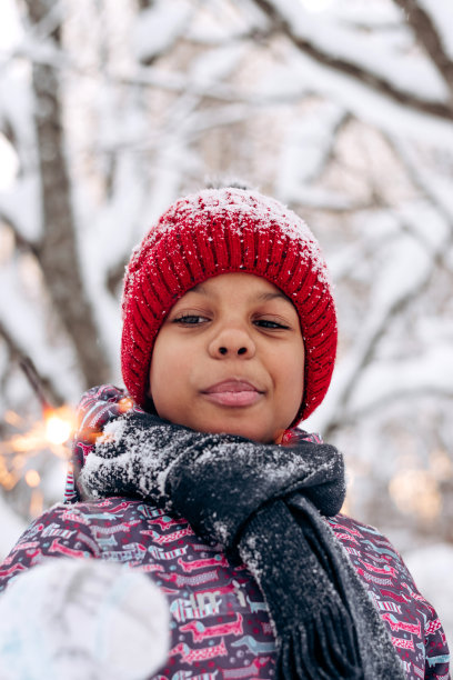 一个快乐的非裔美国小女孩，戴着红帽子，穿着连体衣，手里拿着一个燃烧的火花，在冬天的森林里。冬天的乐趣，圣诞快乐和新年快乐的概念图片下载