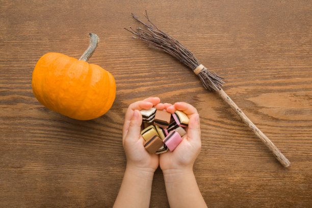彩色果冻软糖和黑巧克力在婴儿张开的手掌。棕色木桌背景是橙色的小南瓜和老女巫扫帚。特写镜头。万圣节饰品。自顶向下的观点。图片下载