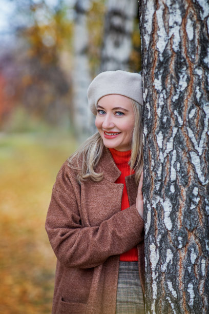 一位戴着贝雷帽、穿着大衣的美丽女子站在一棵秋天的桦树旁微笑着。大自然中人的垂直照片。图片下载